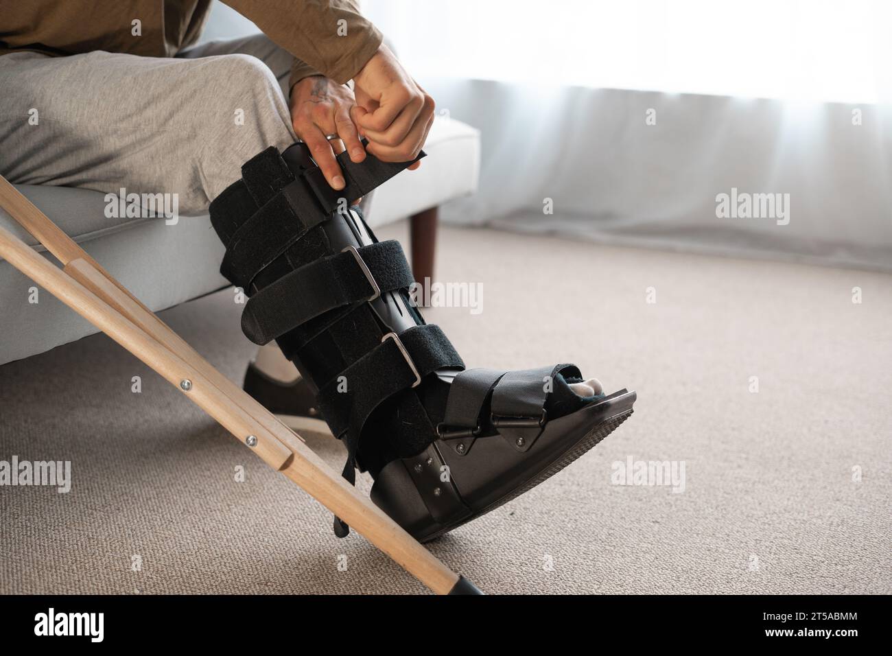 Young man with leg injury using crutch and orthosis sitting on sofa at
