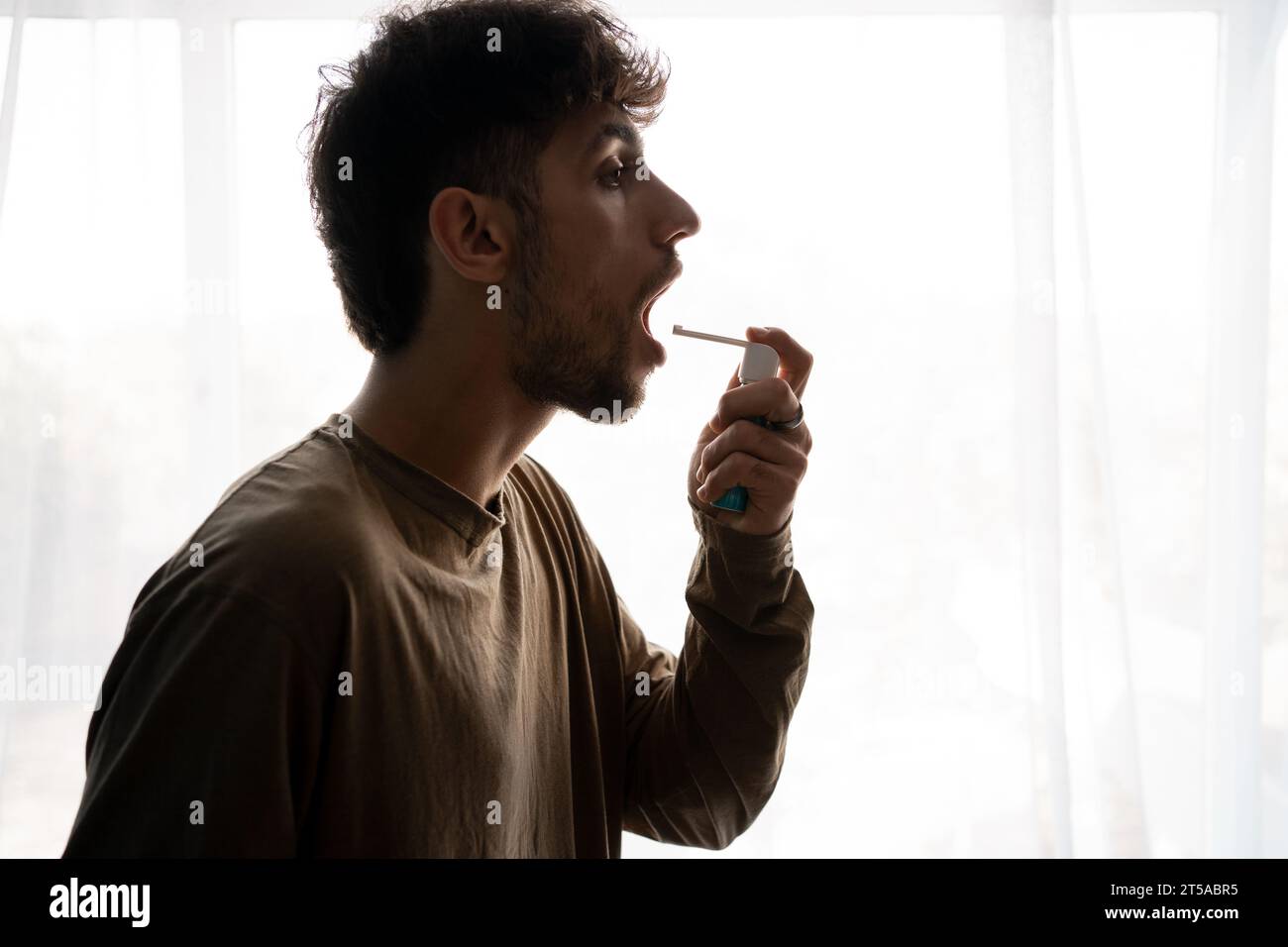 Asthmatic man using an asthma inhaler during asthma attacks standing on window background Stock Photo