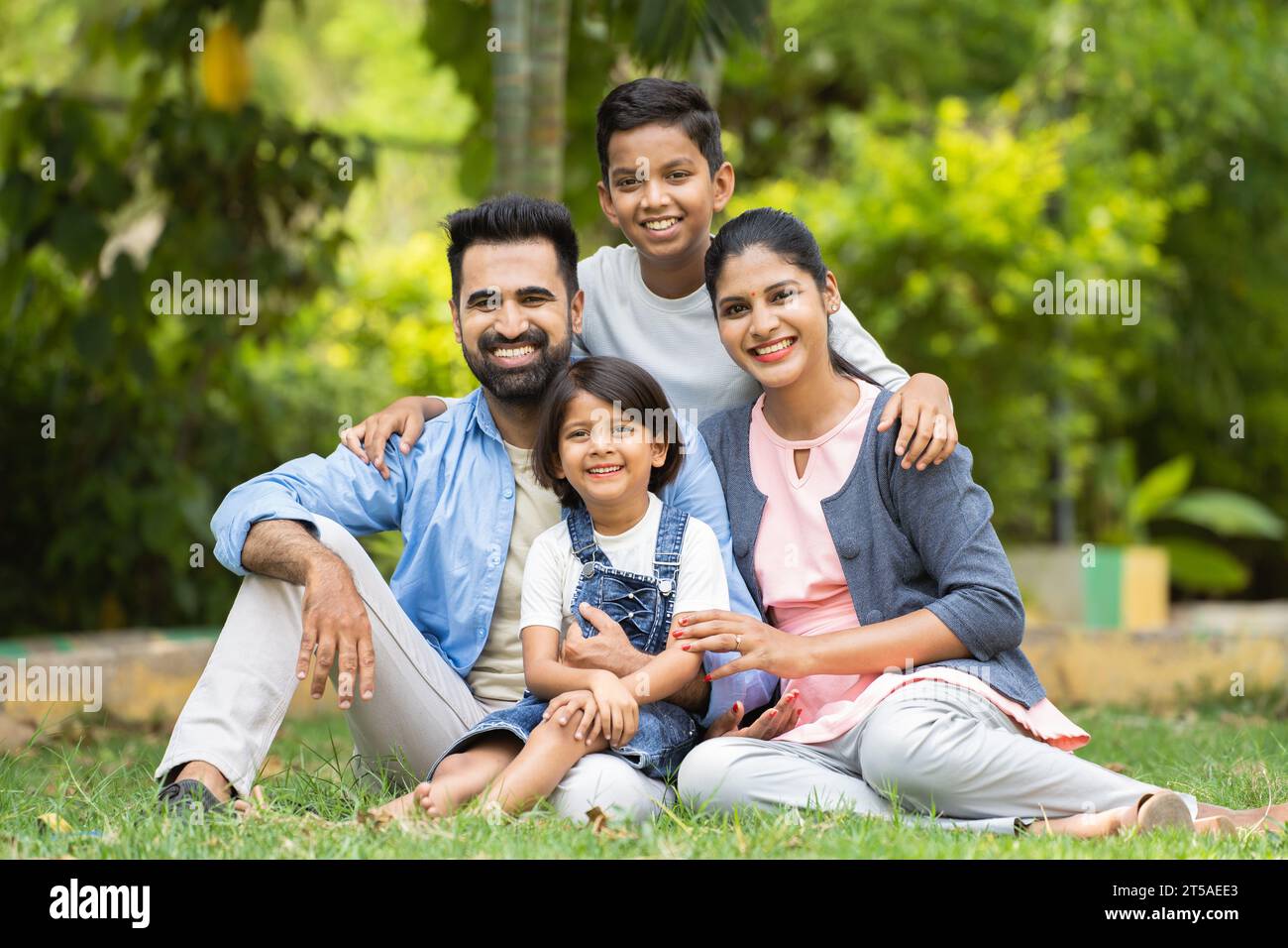 Happy indian couple with kids embracing by looking at camera while sitting at park - concept of relationship, family time and togetherness Stock Photo