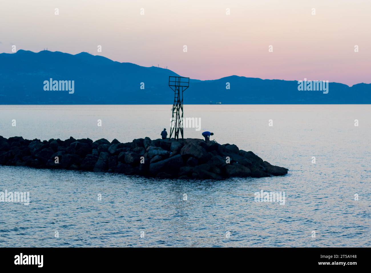 Two fishermen gather their fishing gear at sunset under an old lighthouse. Stock Photo