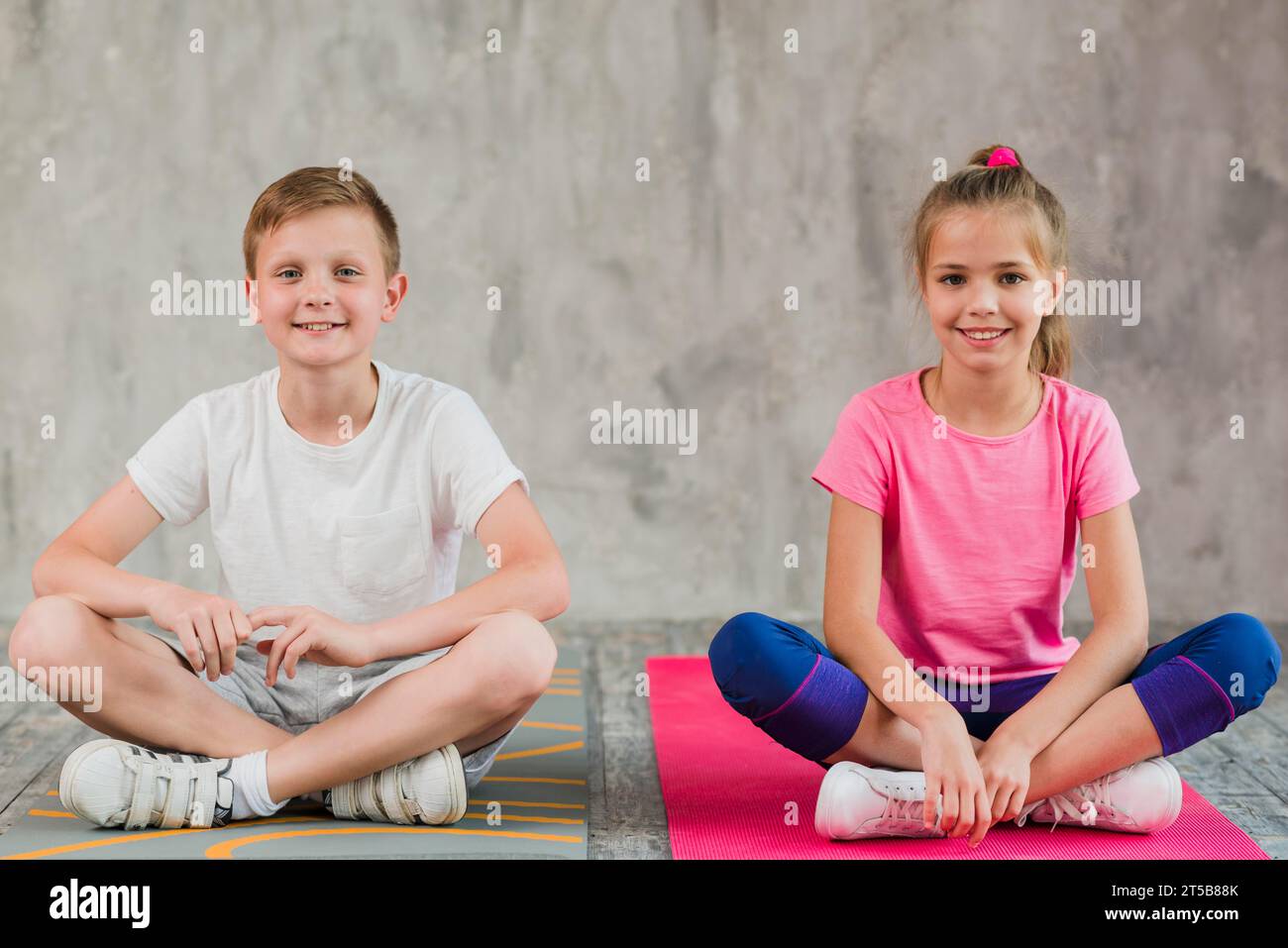 Portrait smiling girl boy sitting exercise mat with their crossed legs front wall Stock Photo