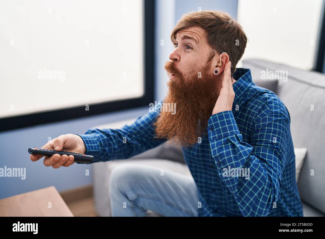 Young redhead man using deafness aid machine watching tv at home Stock Photo