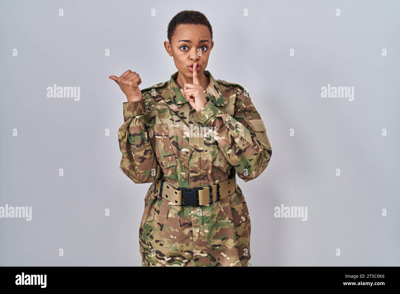 Beautiful african american woman wearing camouflage army uniform asking to be quiet with finger on lips pointing with hand to the side. silence and se Stock Photo