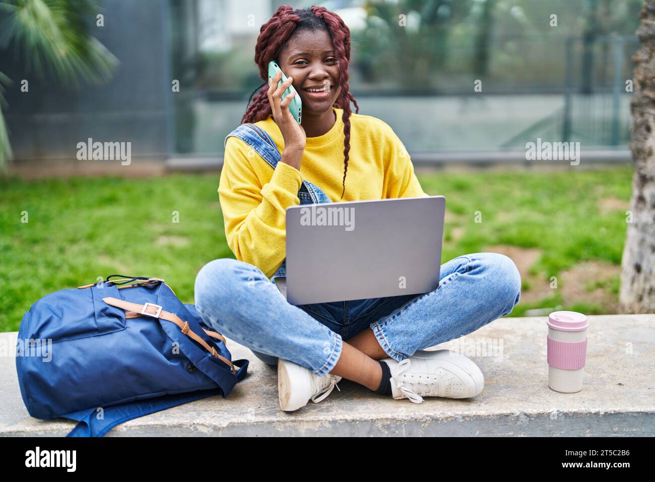 African american woman student using laptop talking on smartphone at ...