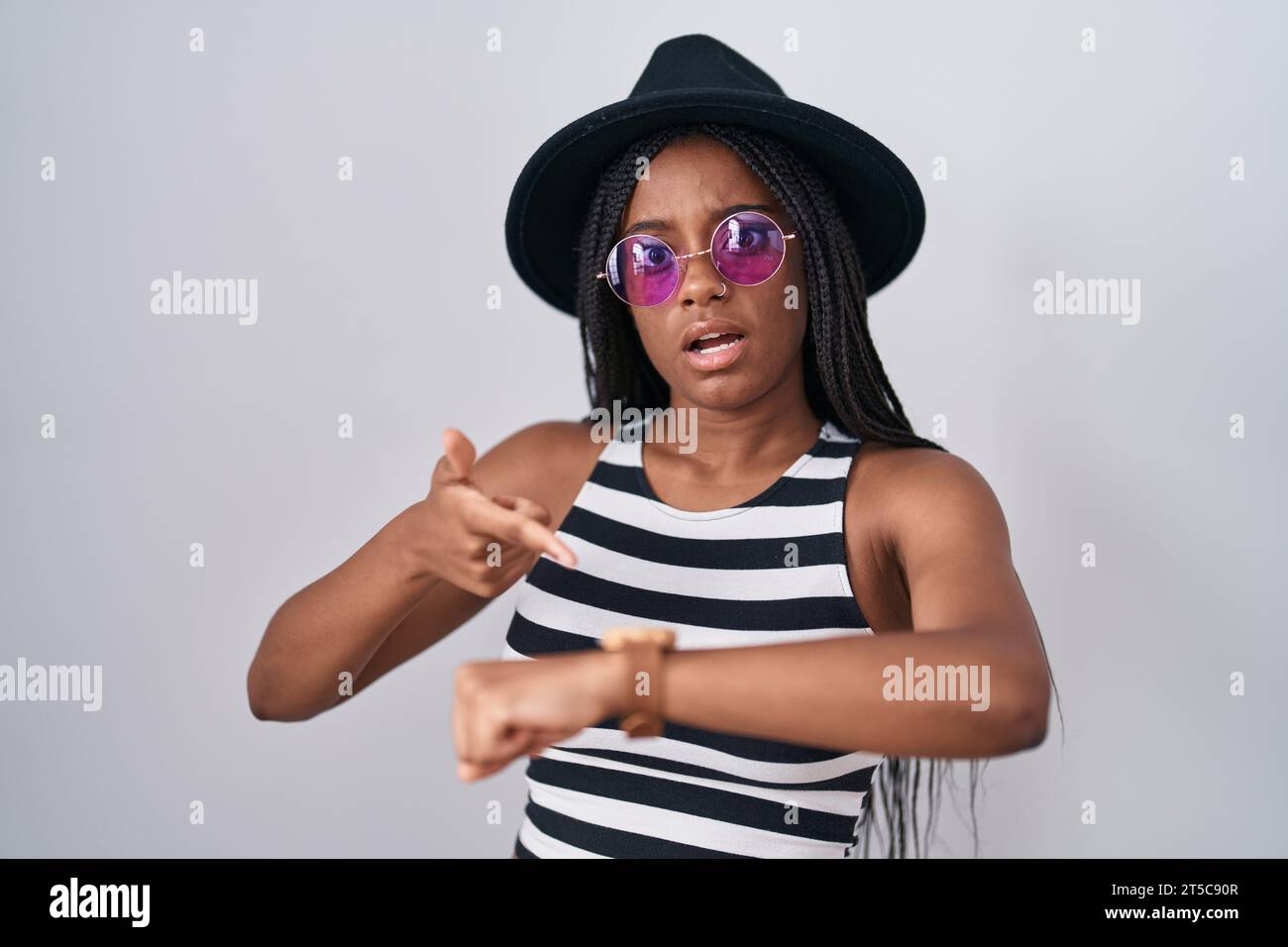 Young african american with braids wearing hat and sunglasses in hurry pointing to watch time, impatience, upset and angry for deadline delay Stock Photo