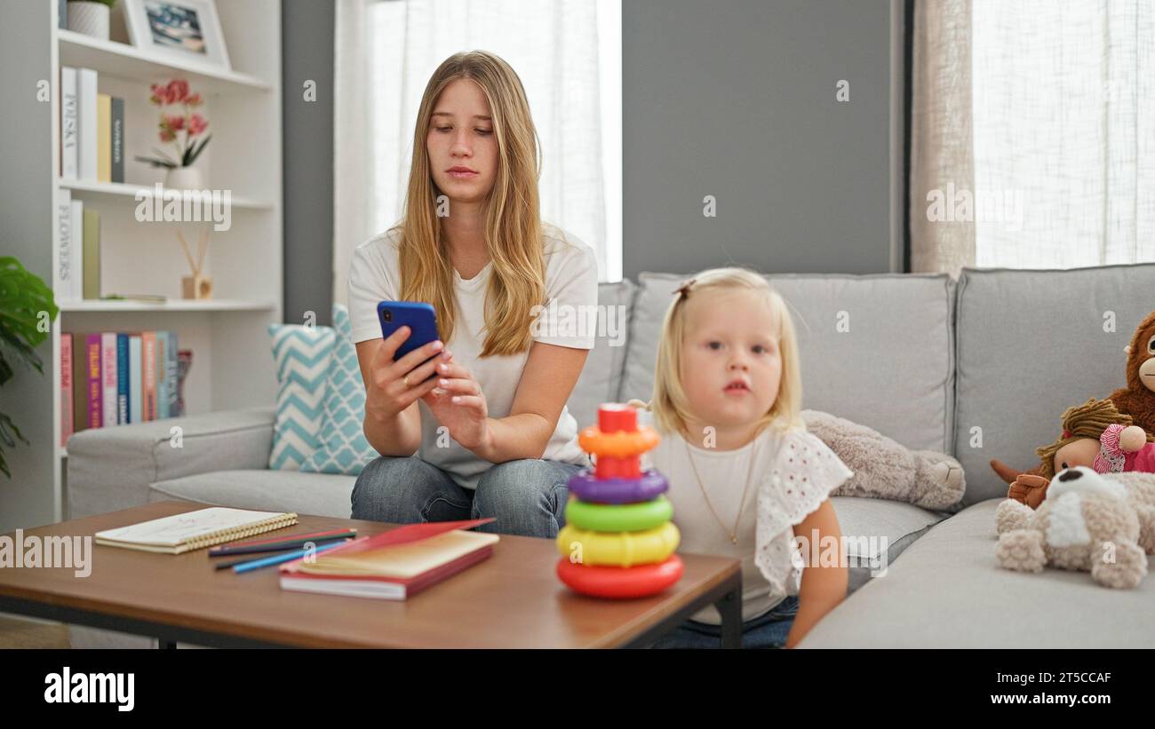 Relaxed caucasian mum and little daughter comfortably playing hoops game on smartphone, resting together on living room sofa, serious expression typin Stock Photo