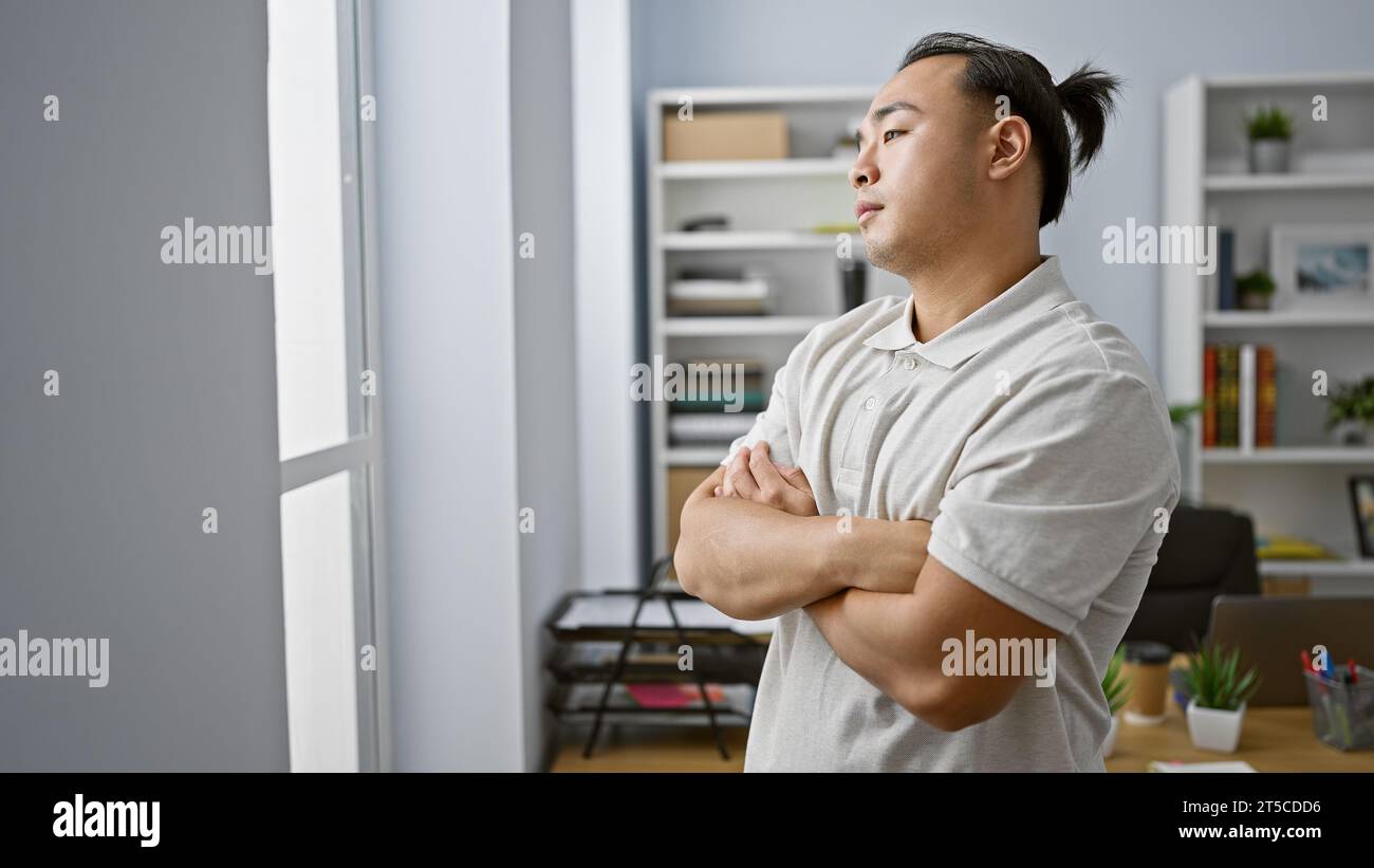 Young chinese man, a focused business worker, throws a side glance out the office window. standing, arms folded, he seems troubled indoors, contemplat Stock Photo