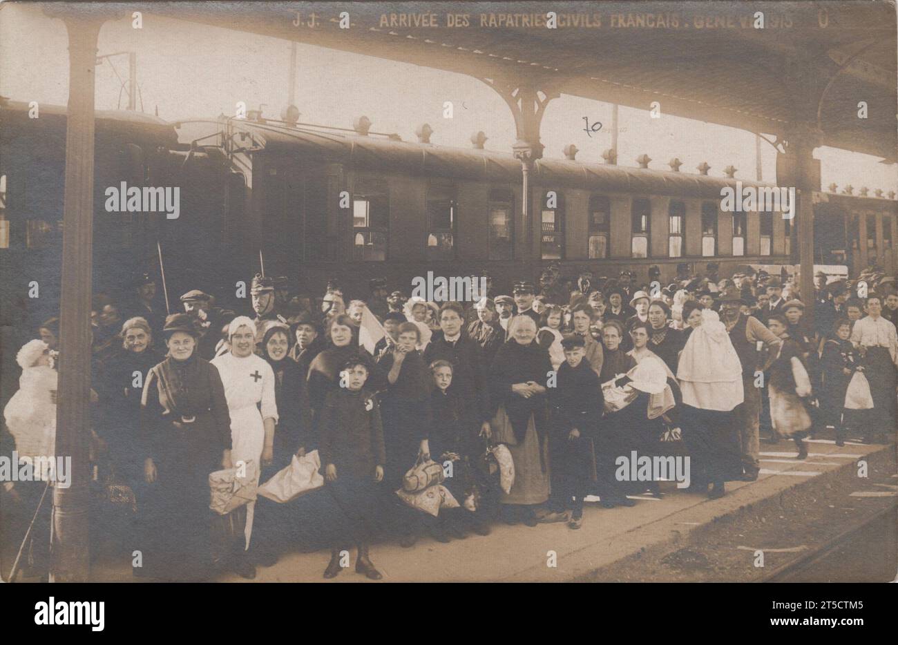 'Arrival of French civilian refugees, Geneva 1915' / 'Arrivée des rapatriés civils Français, Genève 1915': large group of people on a railway station platform, next to a train. Most of the passengers are women and children. A nurse, dressed in white with a red cross on her uniform, is standing towards the front. Some of the people are carrying bundles of belongings. One person is holding a French tricolour flag. Stock Photo