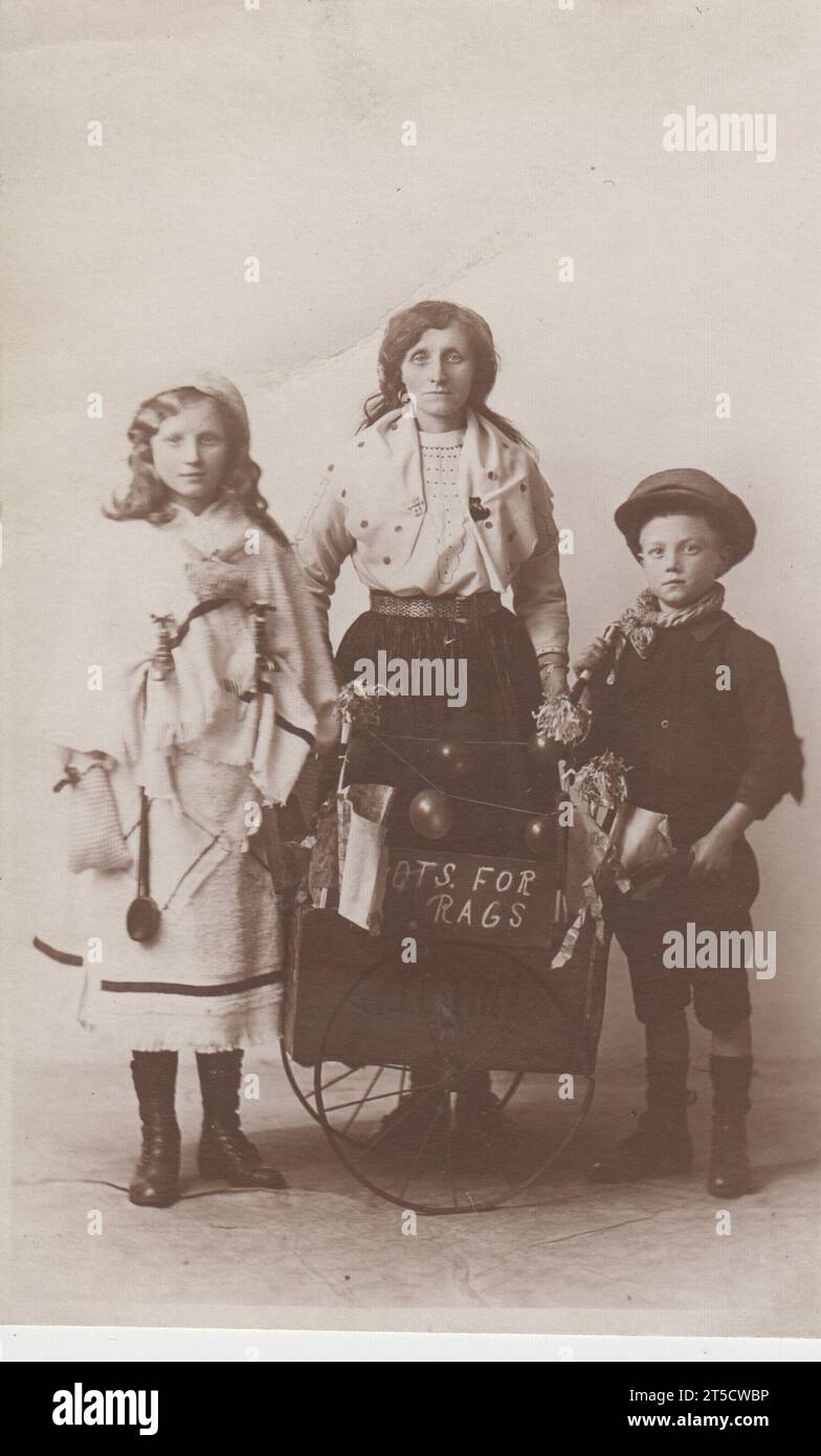 'Pots for rags': early 20th century photograph of a woman and two children standing next to a small handcart which has a sign saying 'Pots for rags' hanging on the side. The girl's outfit incorporates two bathroom / kitchen taps and what appears to be a large metal spoon. The photograph was taken by J.H. Jamieson, a photographer with premises in Preston and Nelson, Lancashire, and Wrexham, North Wales. Stock Photo