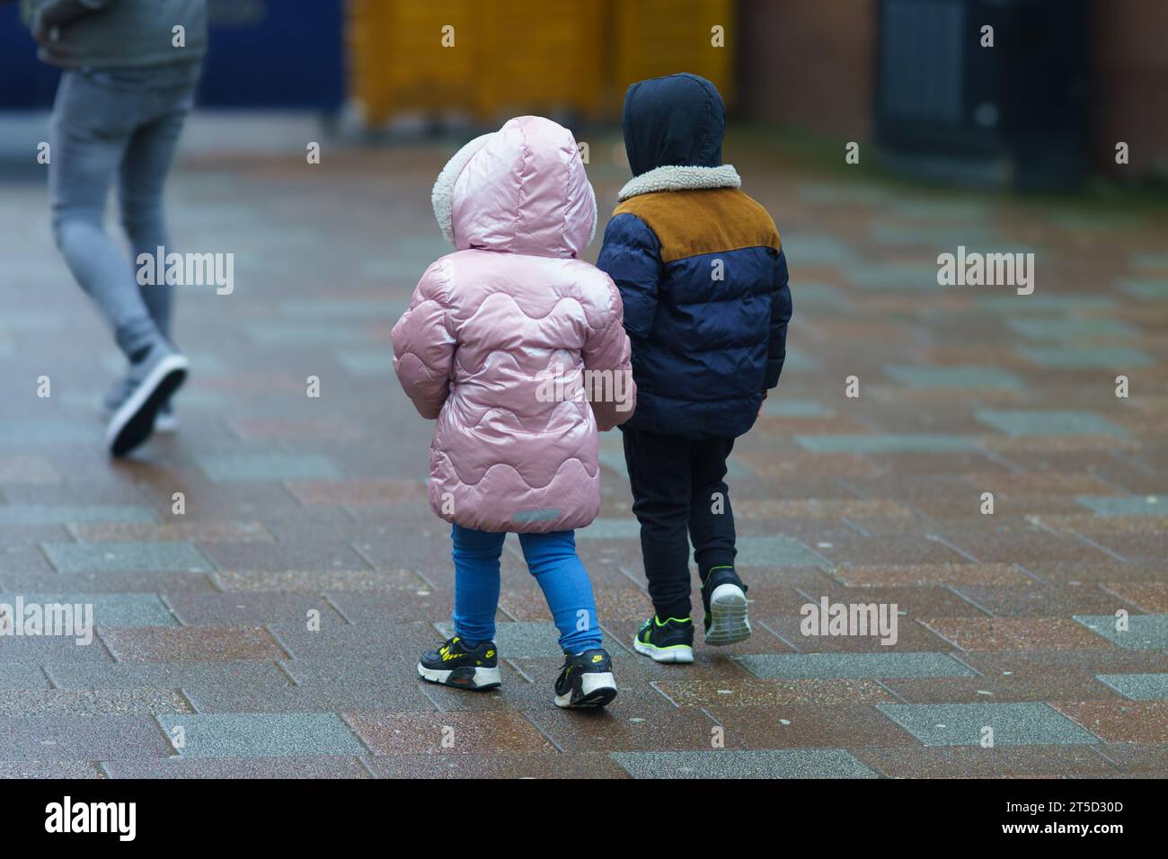 Two young children (a boy and girl) dressed for winter, walk hand in hand behind an adult carer Stock Photo