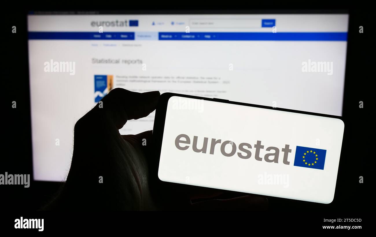 Person holding mobile phone with logo of EU institution European Statistical Office Eurostat in front of web page. Focus on phone display. Stock Photo