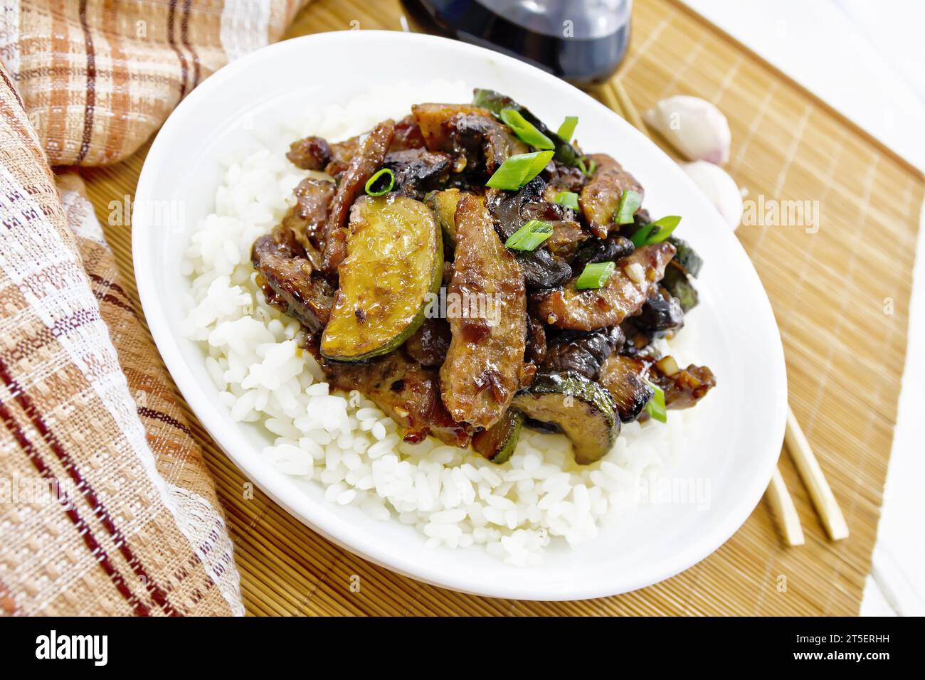Chinese stir-fried dish of chicken meat with zucchini, mushrooms, soy sauce and rice garnish in a plate on bamboo napkin, a towel and garlic on light Stock Photo