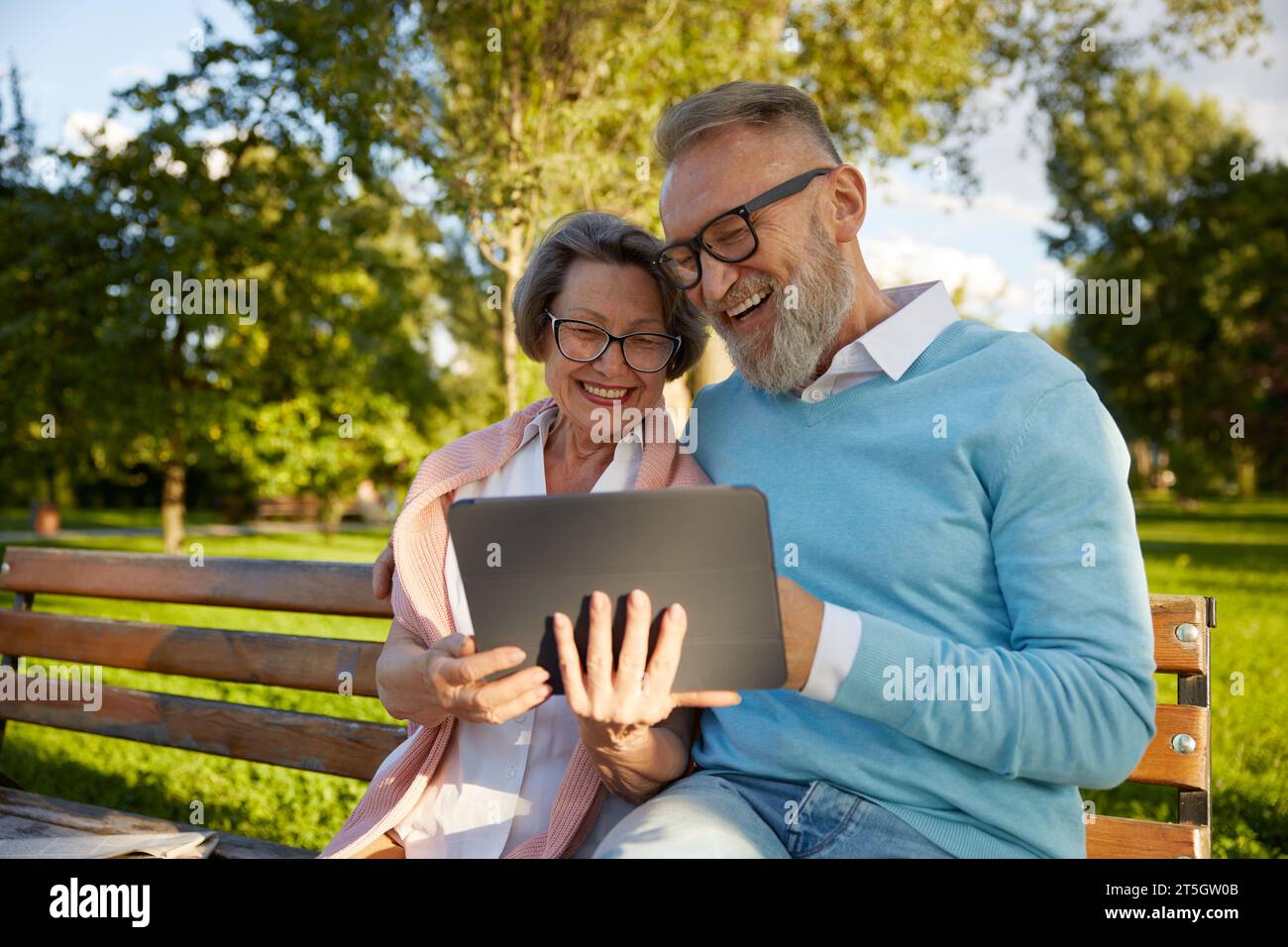 Happy cheerful senior couple using digital tablet while rest on park bench Stock Photo