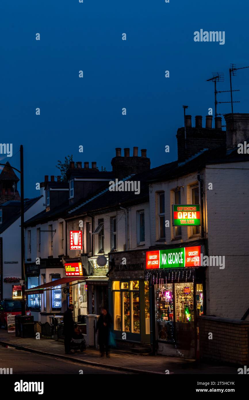 A view along the busy commercial Mill Road Broadway at night. Even at night shops, cafes and bars are brightly lit and open. Cambridge, England, UK Stock Photo