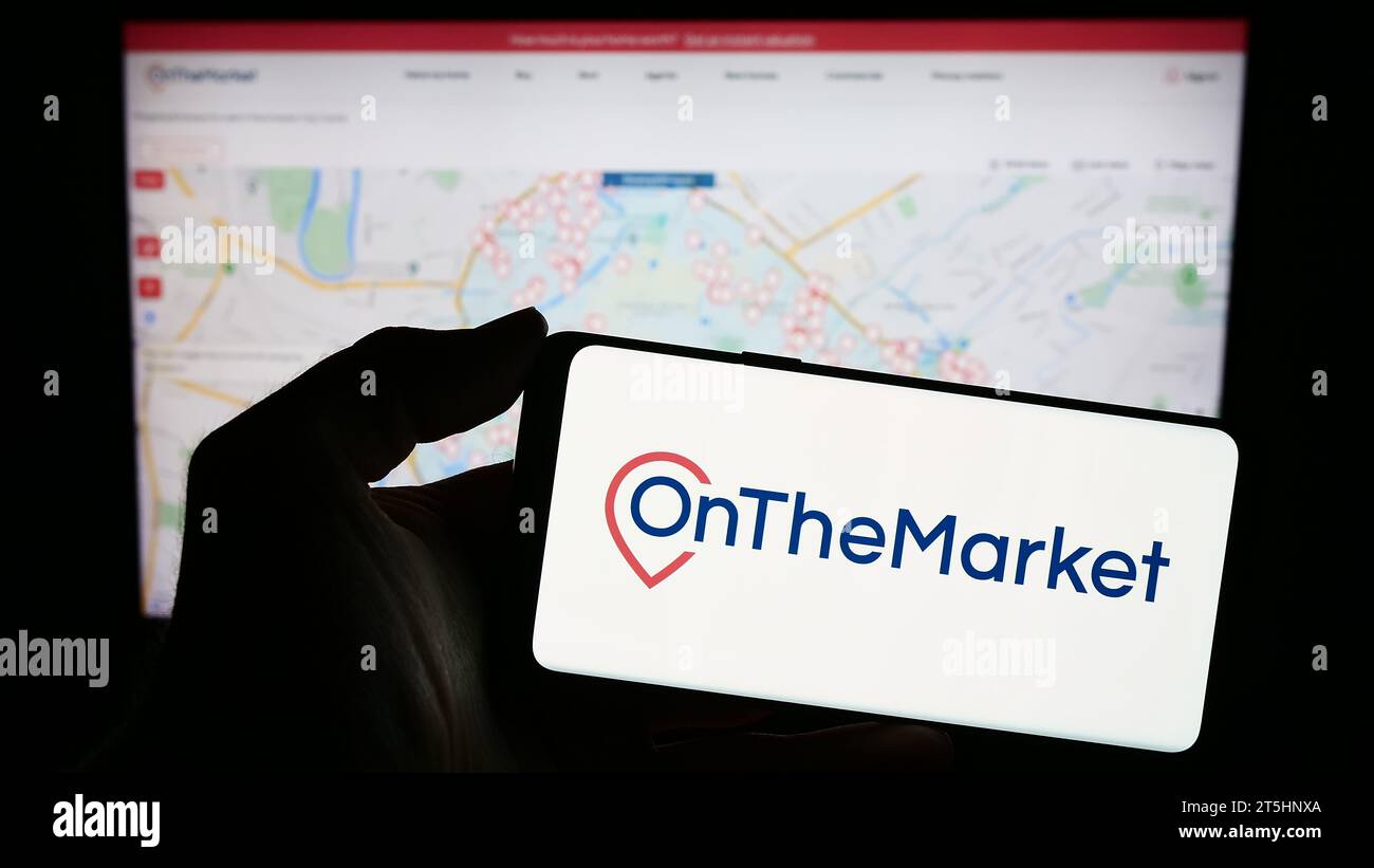 Person holding smartphone with logo of British real estate portal company OnTheMarket.com in front of website. Focus on phone display. Stock Photo