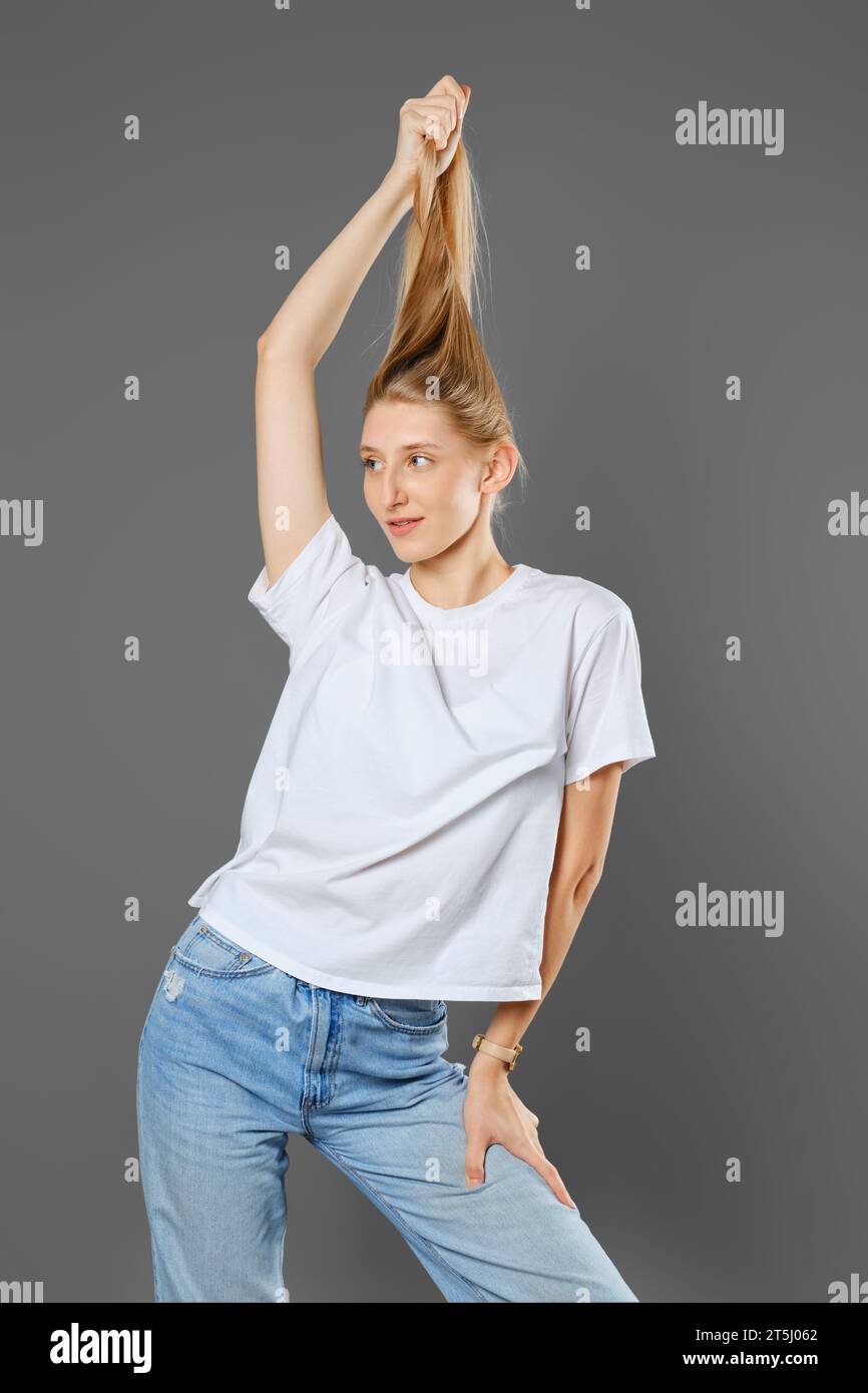 Playful woman wearing blue jeans and white shirt playing with her long blonde hair in studio over grey background Stock Photo