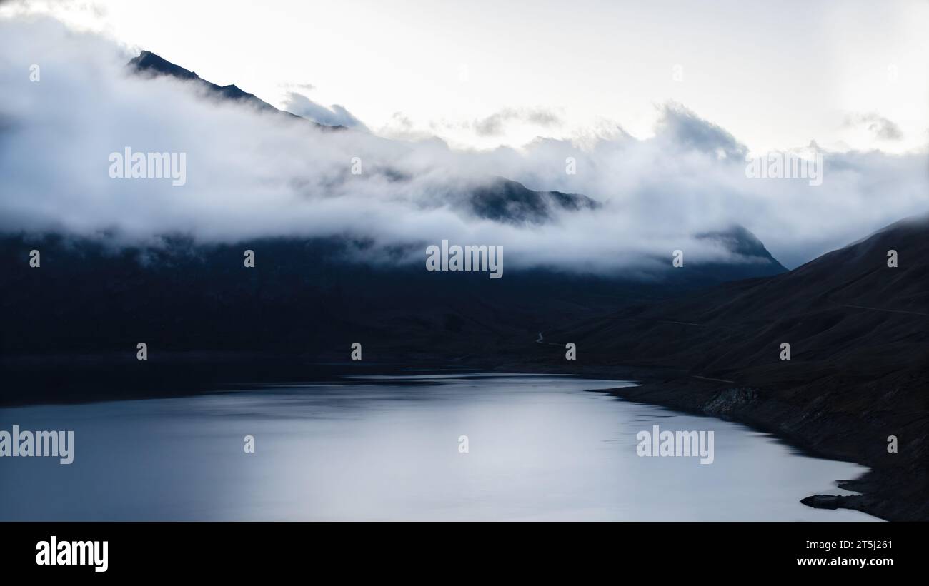 Landscape photohraphy of the Lac du Mont Cenis in the French Alps during the blue hour, dense clouds covering the mountain tops, calm water, dark mood Stock Photo