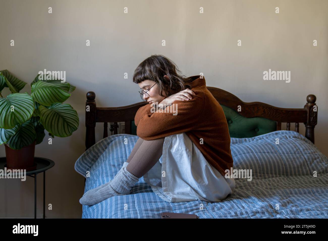 Unhappy depressed teen girl sitting on bed embracing knees in procrastination, apathy and solitude Stock Photo