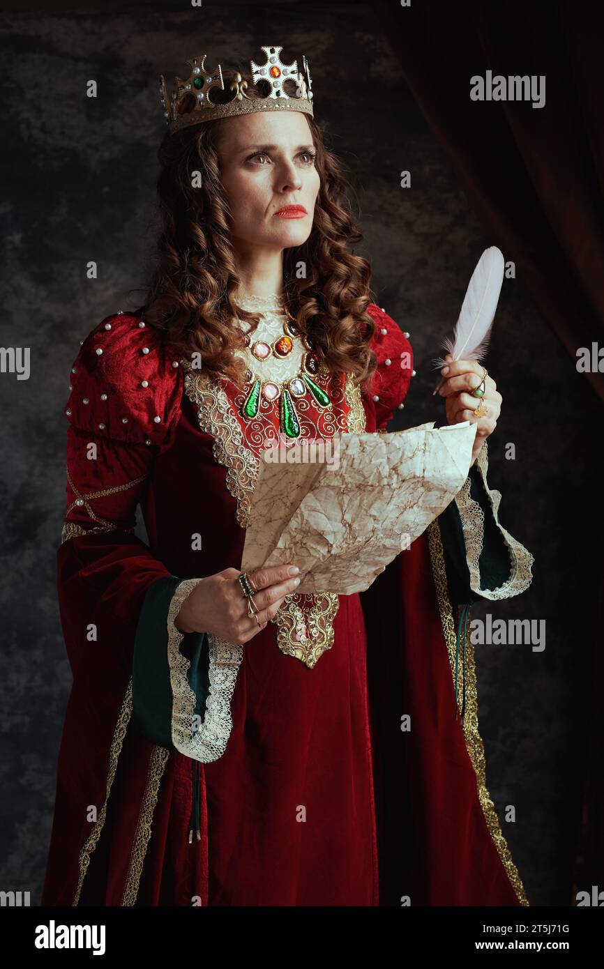 pensive medieval queen in red dress with parchment and crown on dark gray background. Stock Photo
