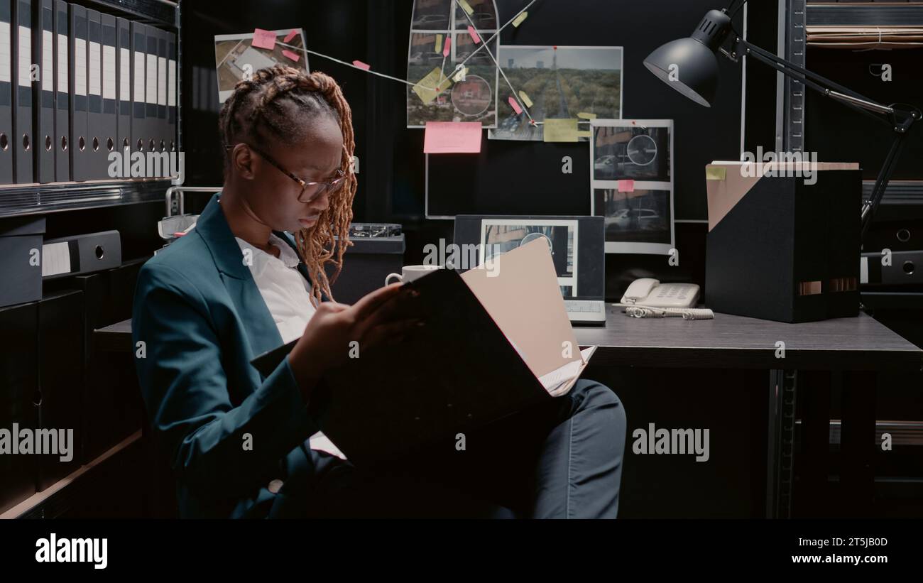 African american policewoman reviewing case files, working on criminal investigation with forensic evidence and suspect photos. Female inspector using detective board in law agency. Stock Photo