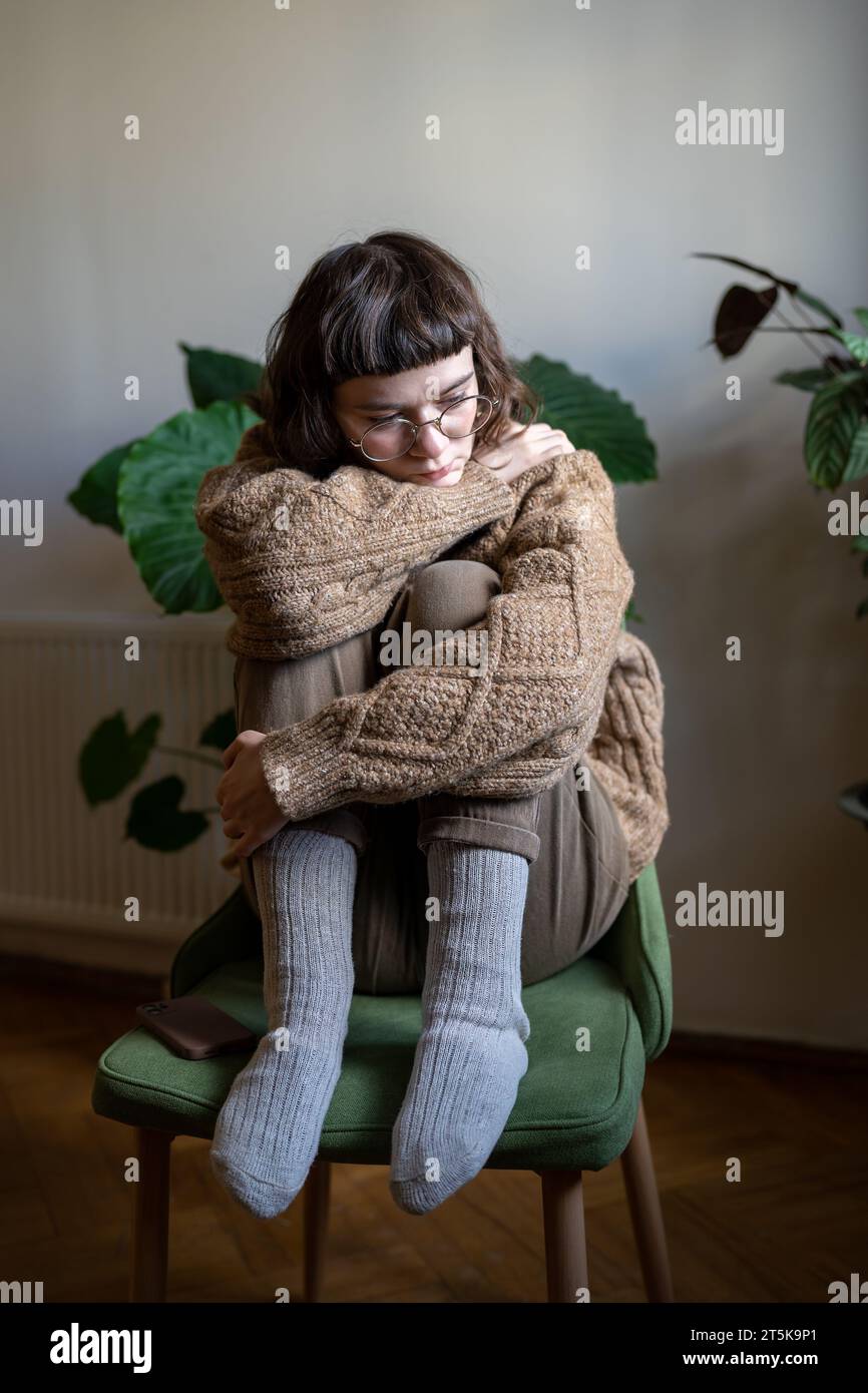Depressed unhappy teen girl sitting alone at home with knees embraced looking indifferently on floor Stock Photo