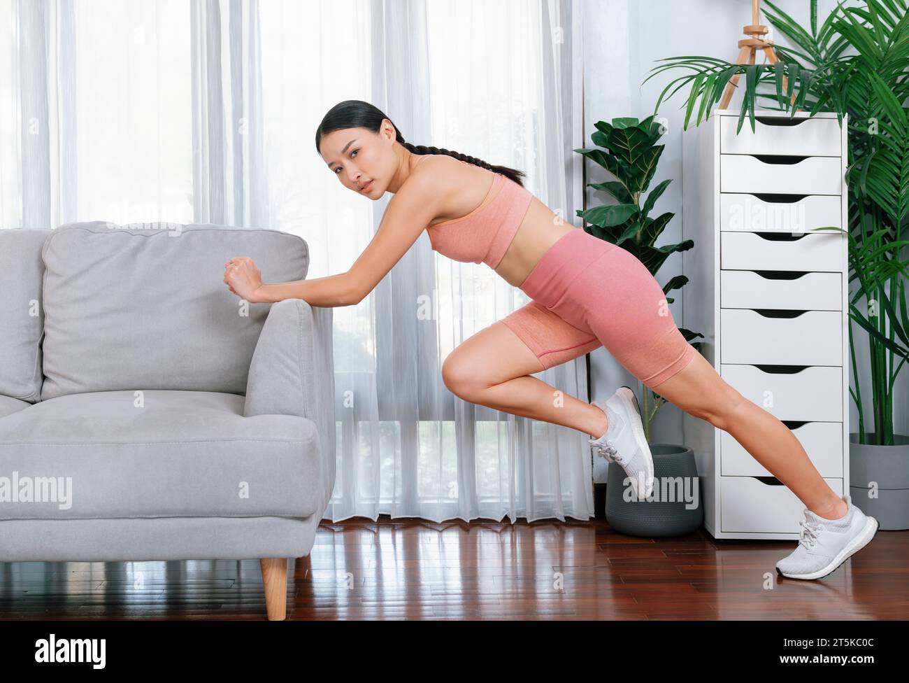 Fit young asian woman stretching her leg her body using sofa in living room. Healthy lifestyle workout training routine at home. Balance and Stock Photo