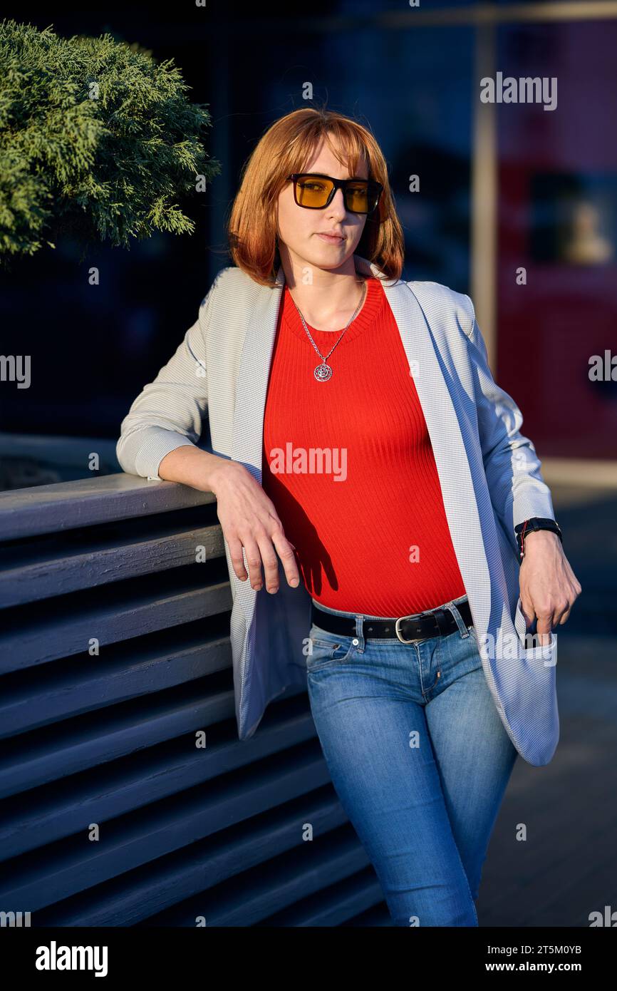 A fashionable redhead woman enjoying an autumn evening in the city. She's wearing a trendy white jacket, a vibrant red t-shirt, and denim jeans, makin Stock Photo