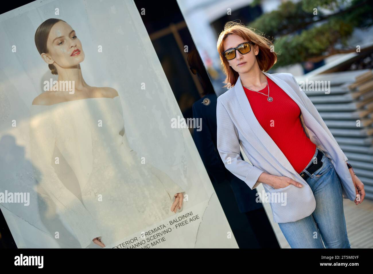 A fashionable redhead woman enjoying an autumn evening in the city. She's wearing a trendy white jacket, a vibrant red t-shirt, and denim jeans, makin Stock Photo