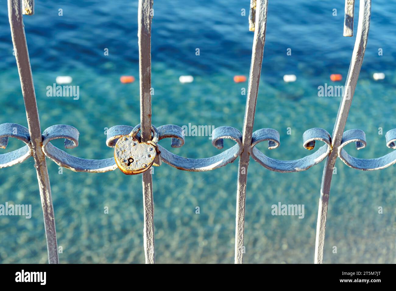 Heart shaped padlock on old metal fence by transparent sea bay closeup. Strong love symbol on grid against ocean water on sunny day. Cute artefact Stock Photo