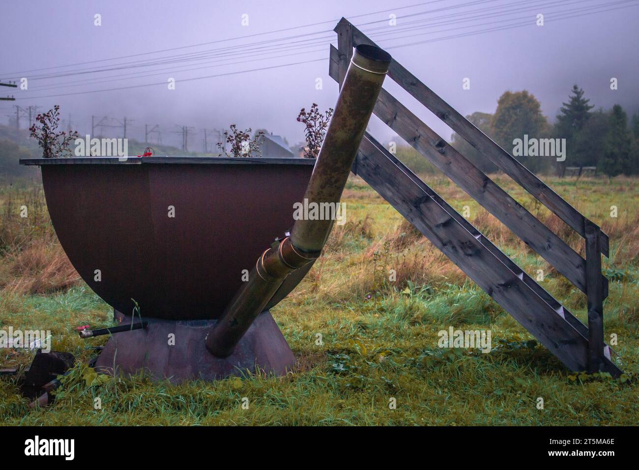 Outdoor bath at foggy back yard. Iron bathtub with wooden stairs on autumn landscape. Empty rusty vat in fall season. Vacations in countryside. Stock Photo