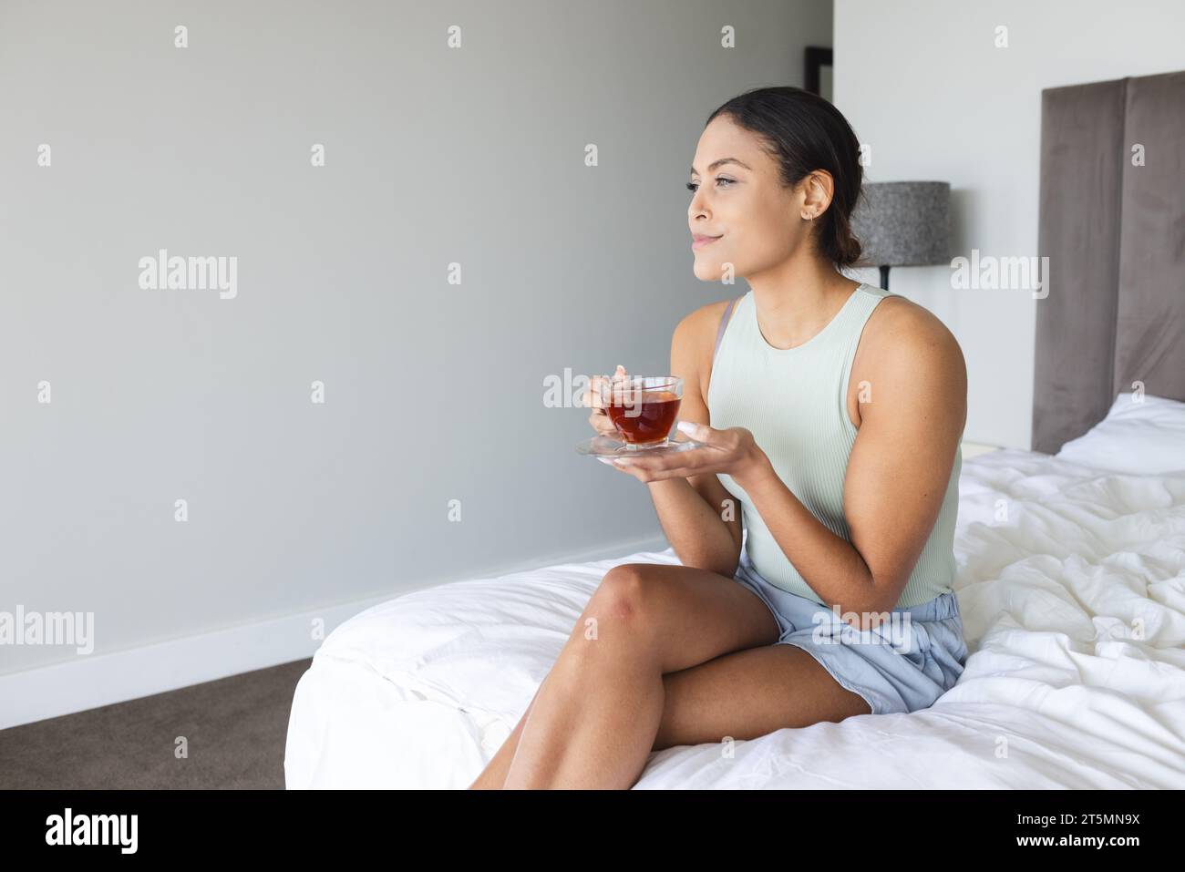 Happy biracial woman sitting and drinking tea in bedroom Stock Photo