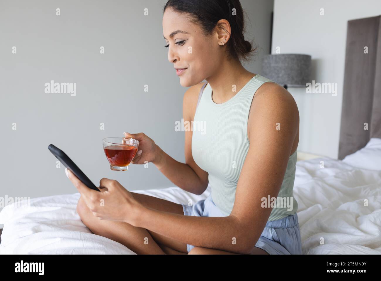 Happy biracial woman using smartphone and drinking tea in bedroom Stock Photo