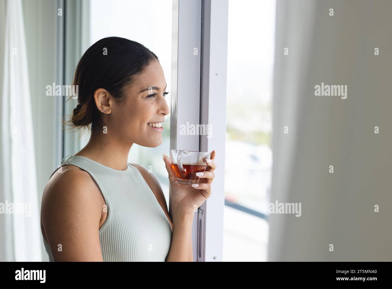 Happy biracial woman at window drinking tea in bedroom Stock Photo