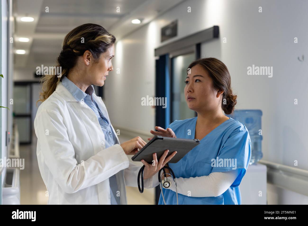 Serious diverse female doctors using tablet discussing in hospital corridor Stock Photo