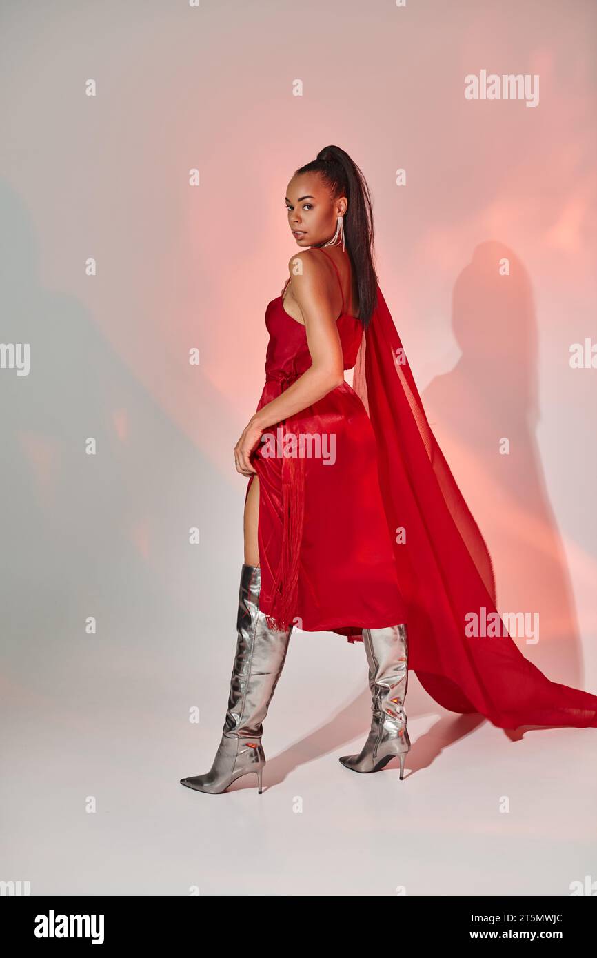 charming african american woman in red dress with shawl posing in silver boots on grey with lighting Stock Photo