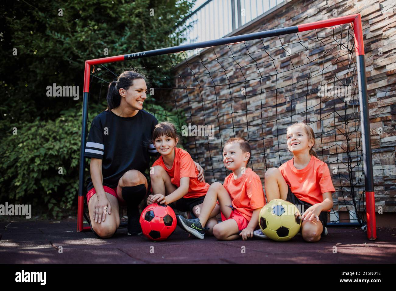 Mom playing football with her children, dressed in football jerseys. The family as one soccer team. Family sports activities outside in the backyard Stock Photo