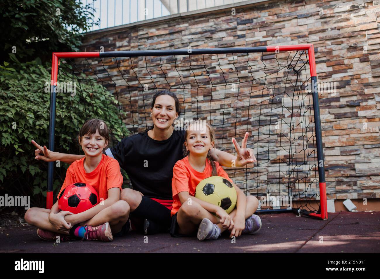 Mom playing football with her daughters, dressed in football jerseys. The family as one soccer team. Family sports activities outside in the backyard Stock Photo