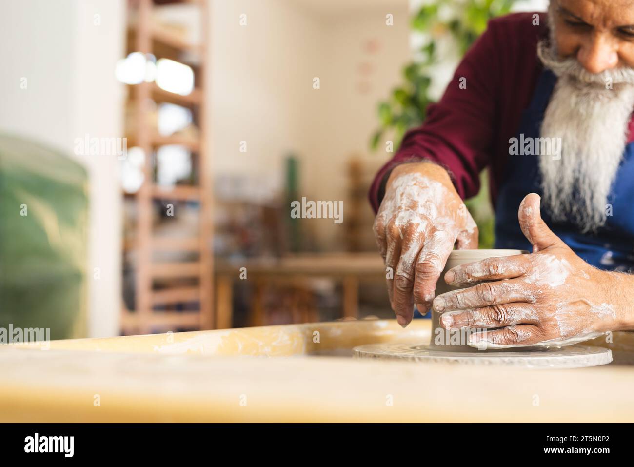 Focused biracial senior potter with long beard using potter's wheel in pottery studio Stock Photo