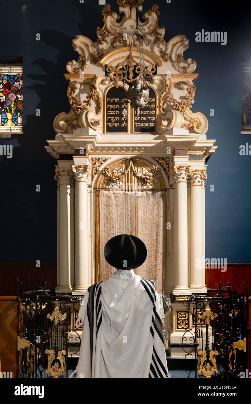 A Jewish man wearing a tallit or prayer shawl leads the synagogue in shacharit morning prayers beneath a decoration of two tablets containing the word Stock Photo