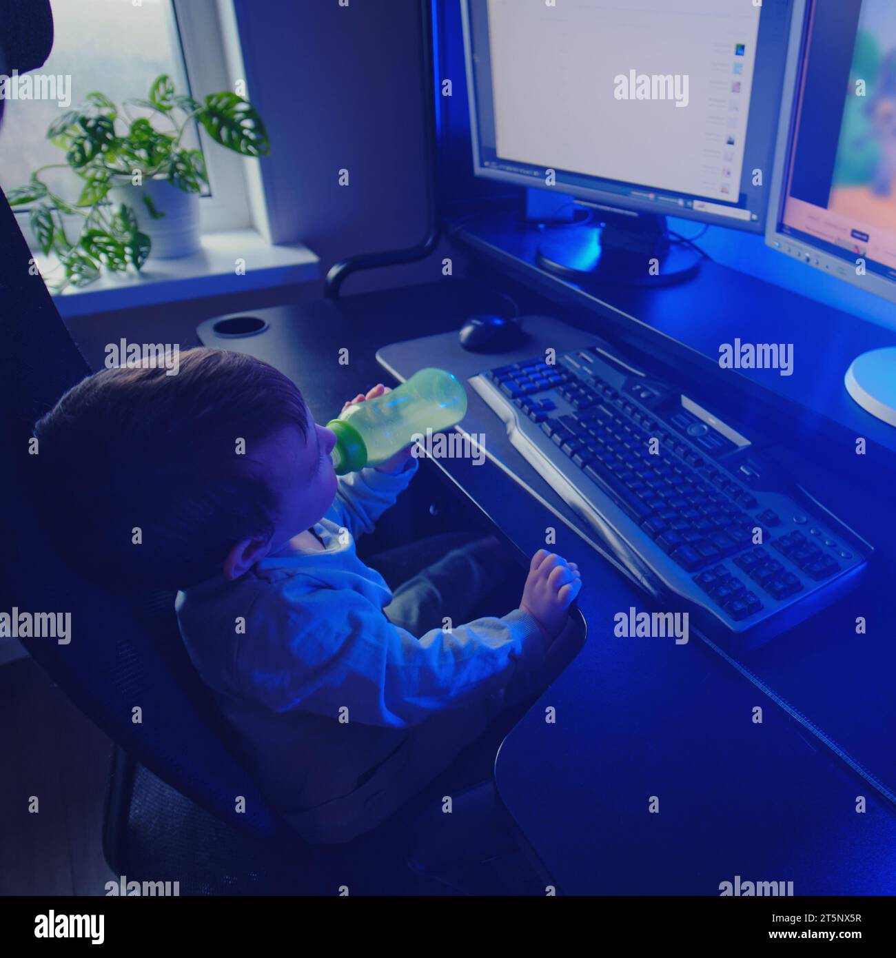 Happy baby is sitting at the computer keyboard with a bottle of milk in his hands. A child with a desktop PC in a home living room drinks milk formula Stock Photo