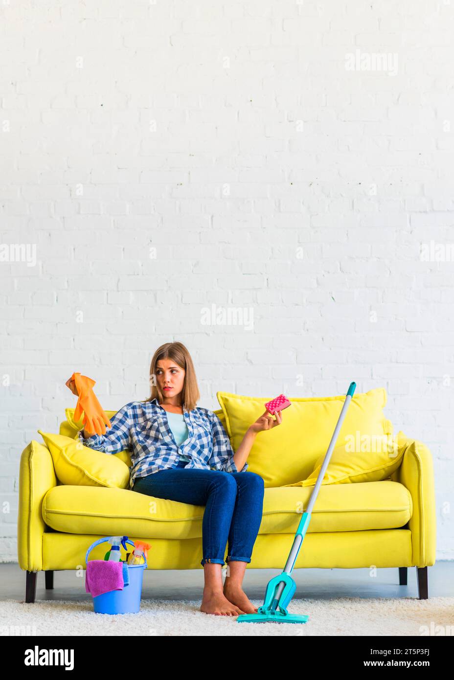 Confused young woman sitting yellow sofa holding gloves brush Stock Photo