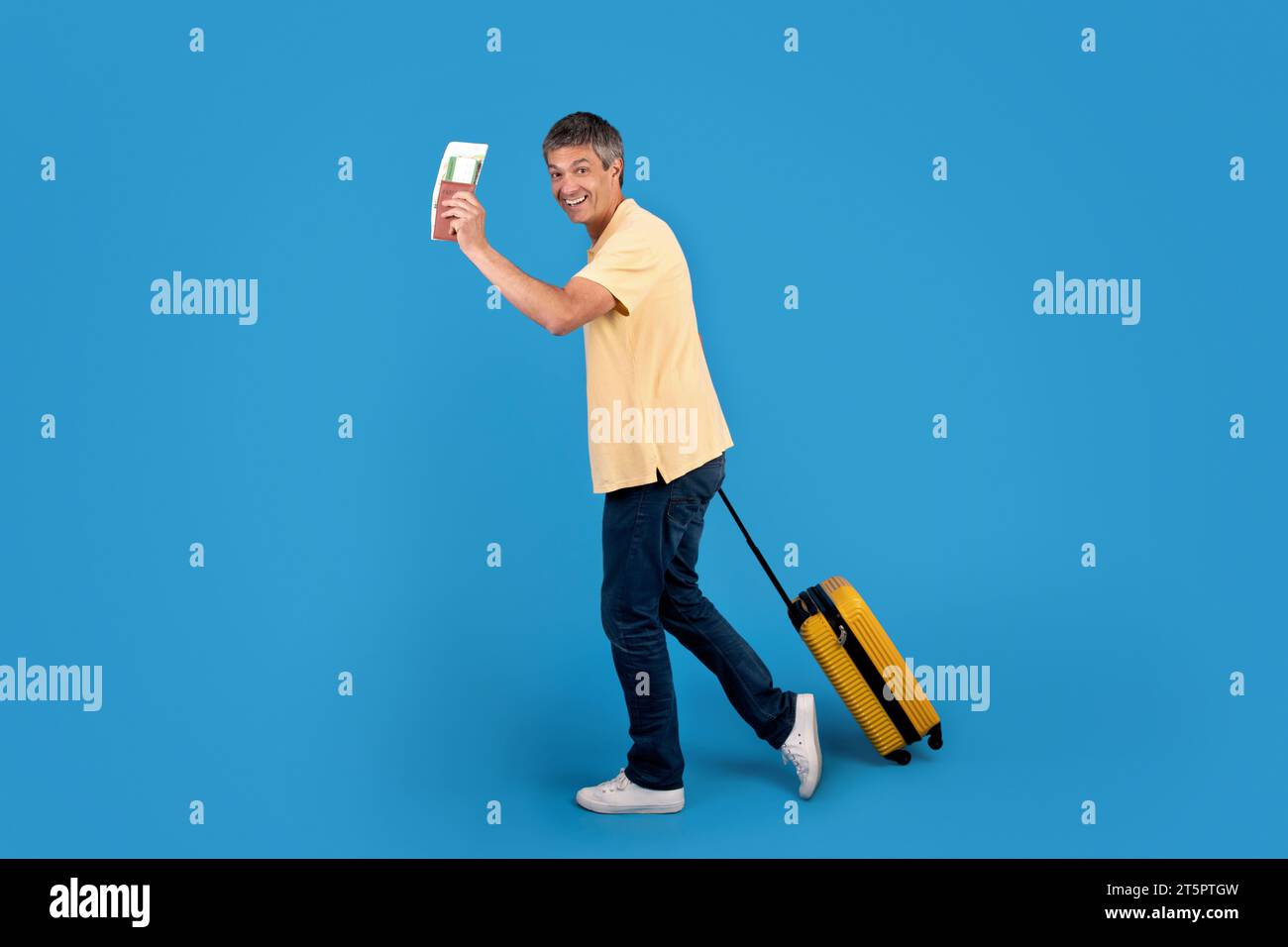 Mature traveler holds boarding pass posing with suitcase, blue background Stock Photo