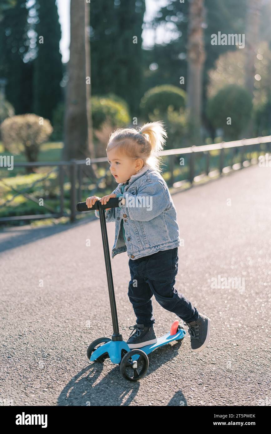 Little girl rides a threewheeled scooter on a road in the park