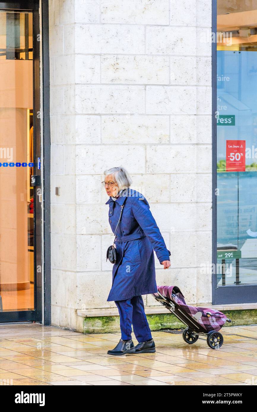 Elderly woman pulling shopping trolley along city pavement in wet weather - Tours, Indre-et-Loire (37), France. Stock Photo