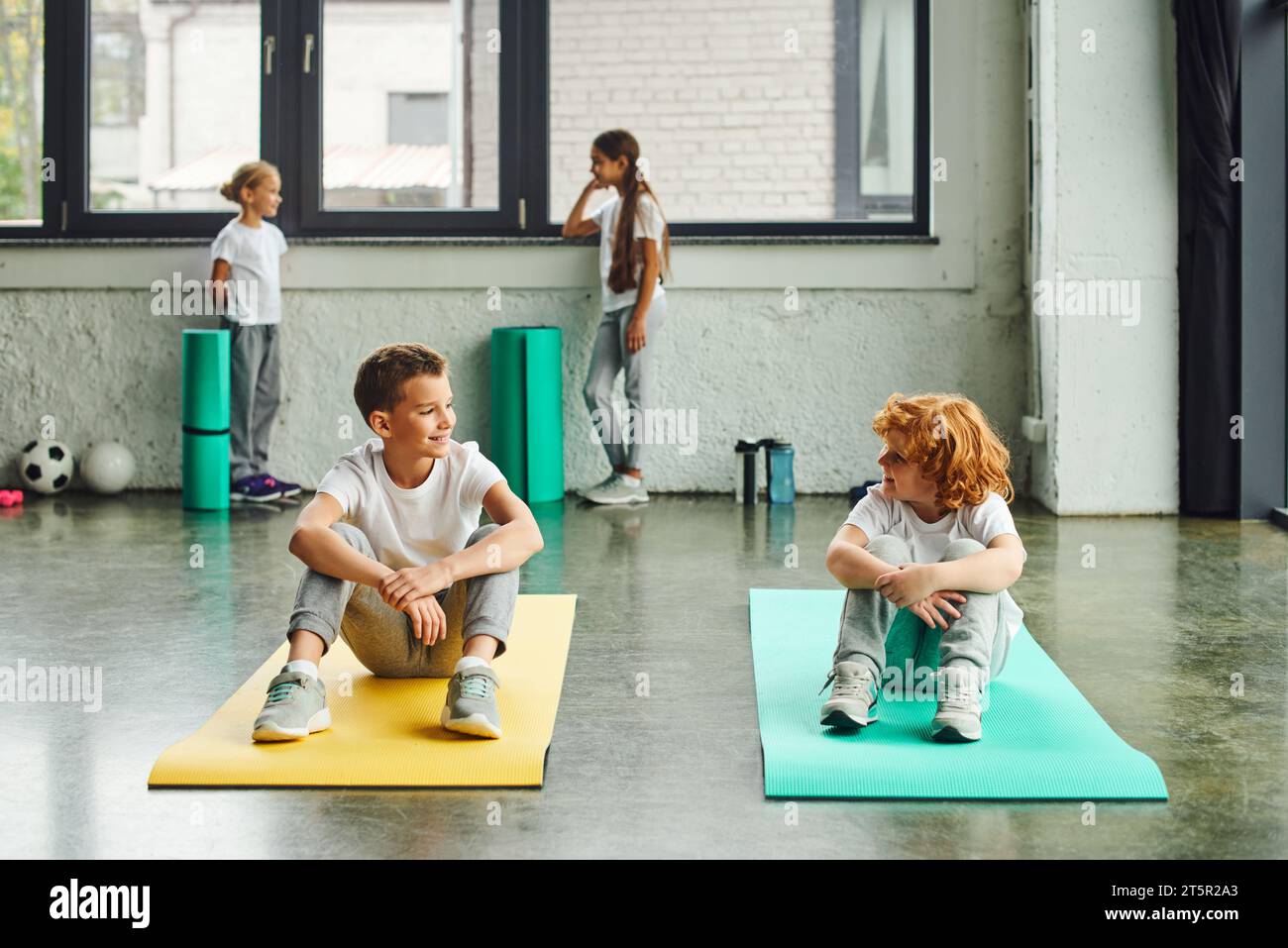 two cheerful boys smiling at each other on fitness mats with cute girls standing on backdrop Stock Photo