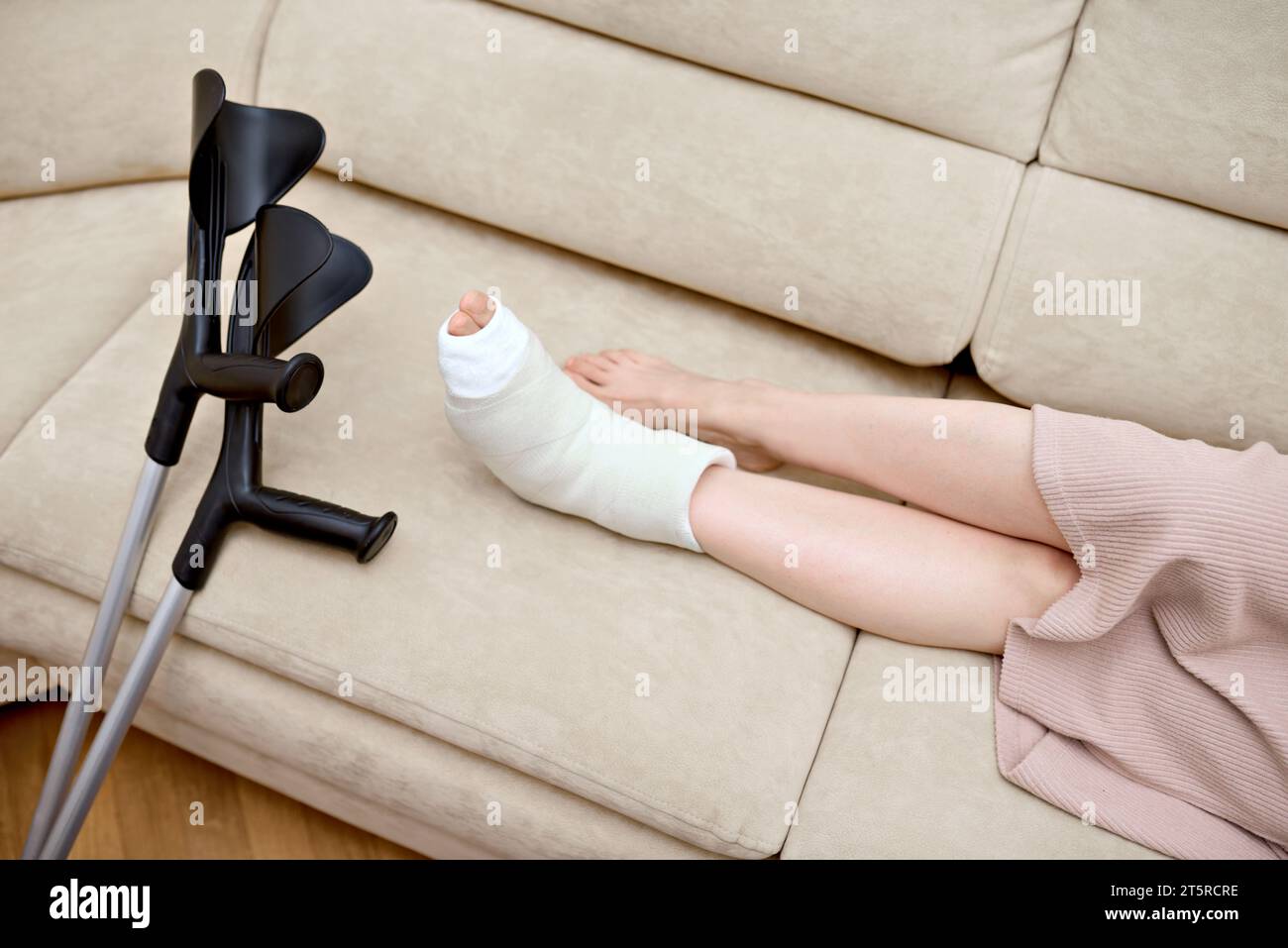 Young woman with foot bone fracture resting on couch at home. Adult's broken leg in a plaster cast. Crutches close by while sitting on the couch Stock Photo