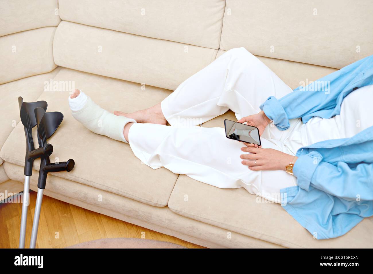 Woman makes a photo of her broken foot for insurance compensation. Female resting on the couch at home with a cast on her fractured leg using a phone. Stock Photo
