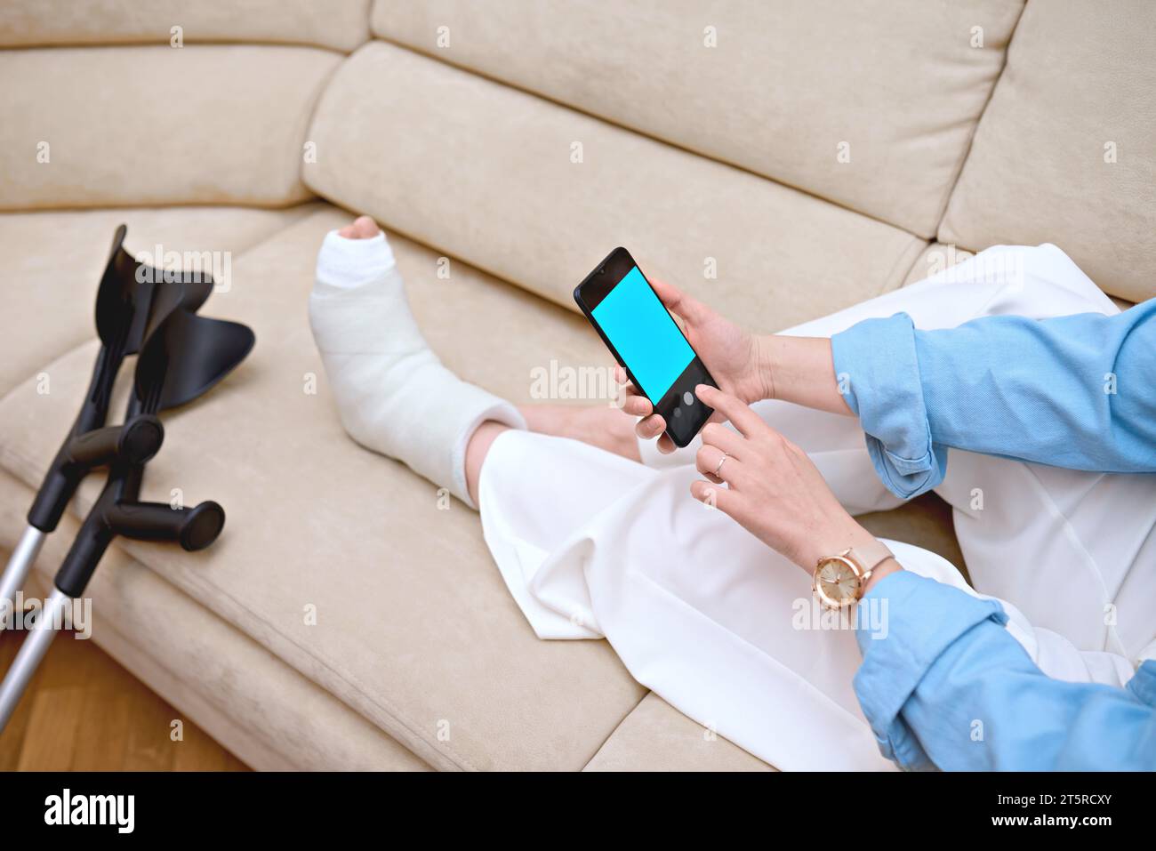 Woman checks possible symptoms with professional traumatologist online via mobile device. A lady, whose leg is in a cast due to an injury Stock Photo