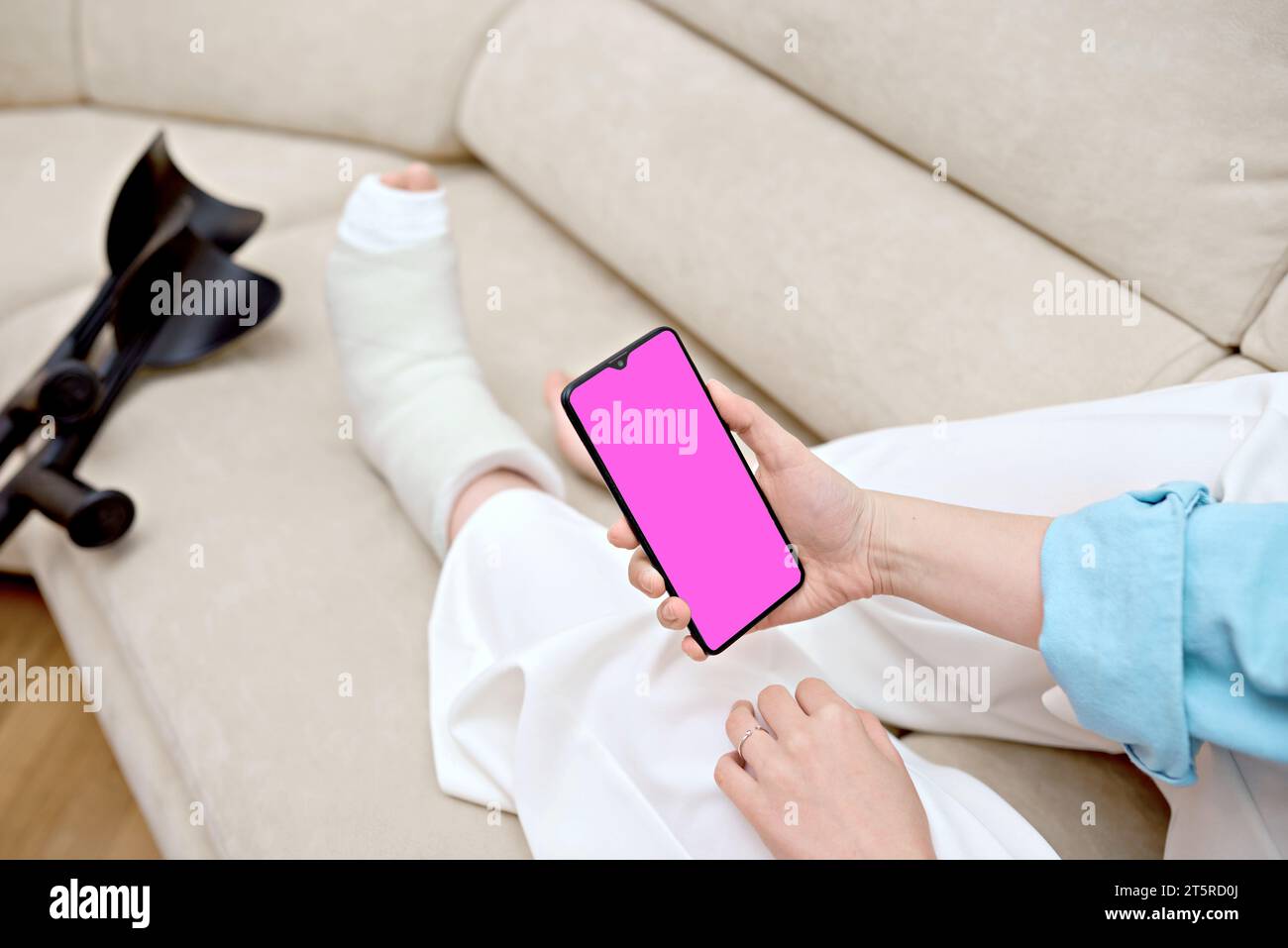 Woman resting on the couch at home with cast on  fractured leg using a phone. Adult checks possible symptoms with professional traumatologist. Stock Photo