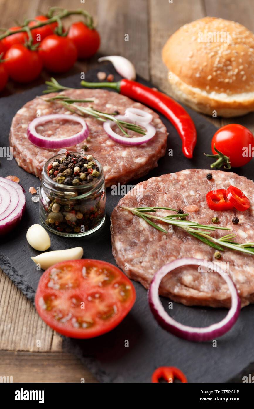 Raw fresh ground beef burger patties, rosemary, vegetables and spices on a black stone plate on a wooden background. Ingredients for a hamburger. Stock Photo